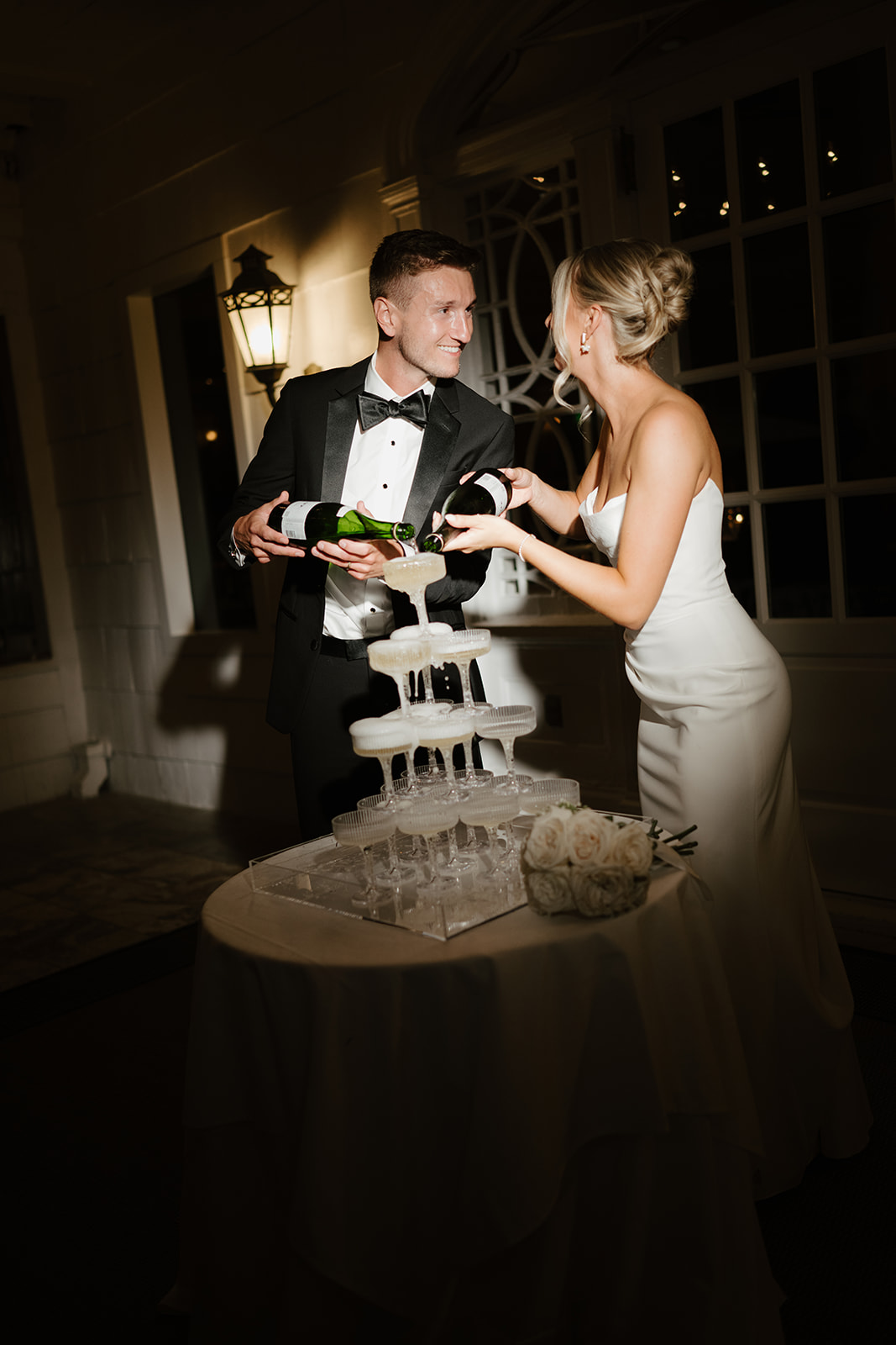 Bride and groom pour champagne into a tiered tower of glasses, laughing together under the night sky.